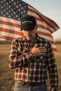 Man wearing the Faith & Grit 1776 • 2026 patriotic hat with hand over heart in front of a waving American flag.