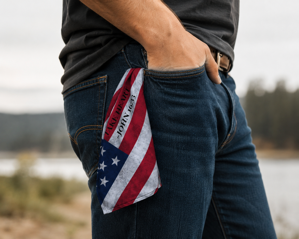American flag inspired Christian bandana with "Take Heart – John 16:33" displayed from a jeans pocket in an outdoor setting, representing faith, strength, and hope through life's challenges.