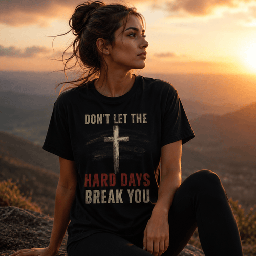 Woman wearing a black Don't Let the Hard Days Break You Christian T-shirt with distressed cross graphic while sitting on a mountainside at sunset. Inspirational faith-based apparel representing resilience, hope, and overcoming life's challenges.