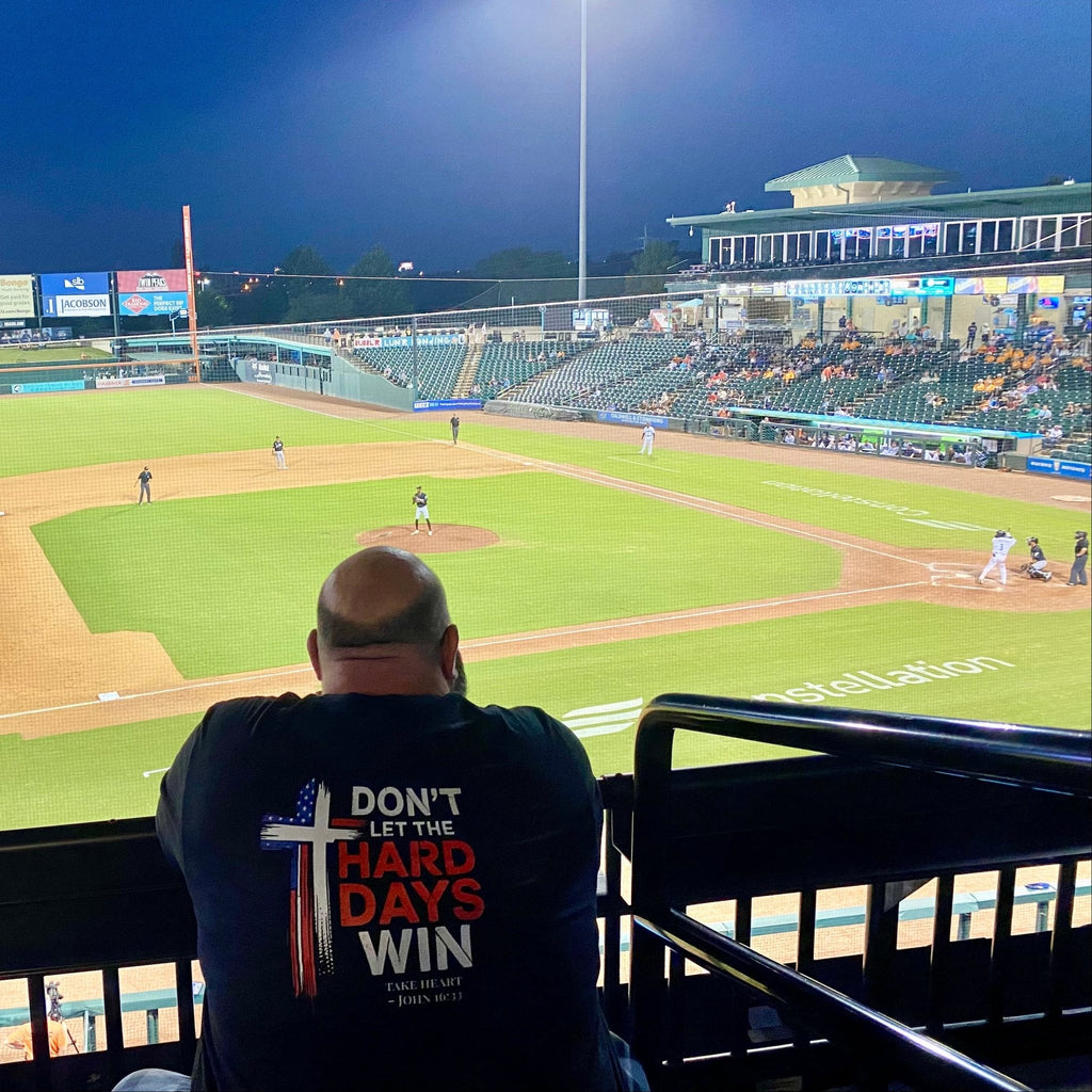 Shane, founder of Held & Anchored, wearing the Don't Let the Hard Days Win Back Print Tee at a baseball game — John 16:33