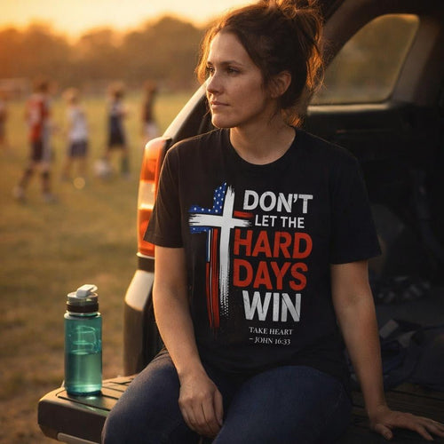 Woman sitting on the tailgate of a pickup truck at sunset wearing the Don't Let the Hard Days Win Christian t-shirt featuring a distressed American flag cross and John 16:33 scripture, reflecting on life's challenges with quiet strength and faith.