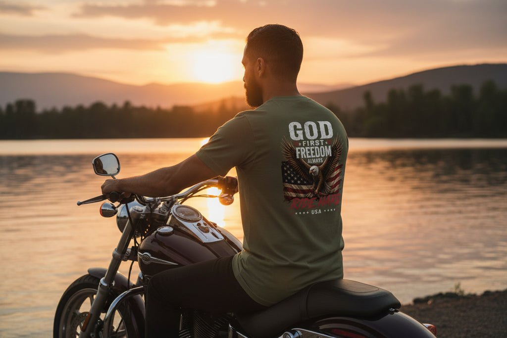 Man sitting on a motorcycle by a lake at sunset, wearing a t-shirt with text and graphics.