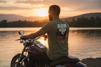 Man sitting on a motorcycle by a lake at sunset, wearing a t-shirt with text and graphics.