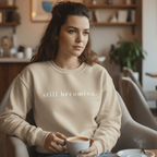 Woman sitting in a cozy cafe holding a cup of coffee, wearing a beige sweatshirt with text.