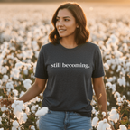Woman standing in a field of cotton wearing a t-shirt with 'still becoming'.