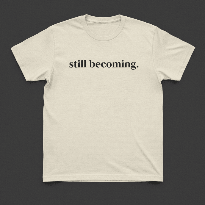Beige t-shirt with 'still becoming.' text on a dark gray background