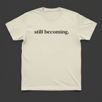 Beige t-shirt with 'still becoming.' text on a dark gray background