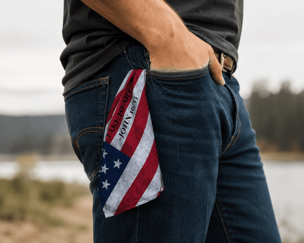 American flag inspired Christian bandana with "Take Heart – John 16:33" displayed from a jeans pocket in an outdoor setting, representing faith, strength, and hope through life's challenges.