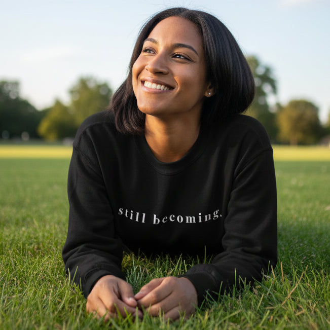 Person wearing a black sweatshirt with 'still becoming.' while laying in a field and smiling. 