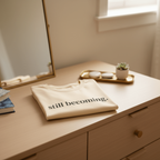 Folded beige sweatshirt with 'still becoming' text on a wooden surface.